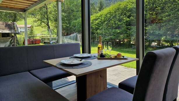 Een moderne eethoek buiten met een houten tafel en grijs gestoffeerde zitplaatsen, met uitzicht op een weelderig groen landschap en een berg in de verte.