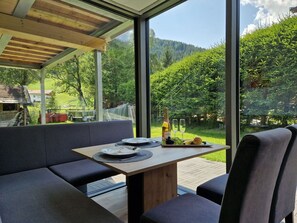 Un área de comedor al aire libre moderna con mesa de madera y asientos acolchados grises, con vistas a un paisaje verde exuberante y una montaña en la distancia.