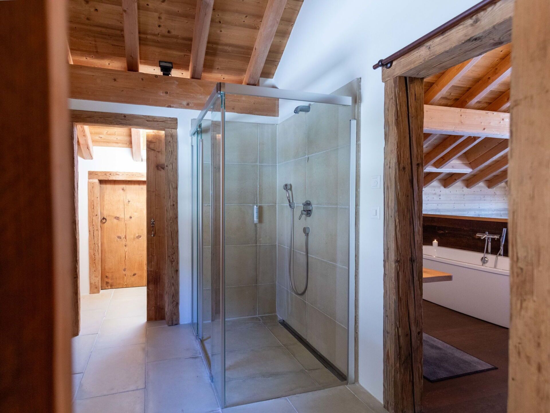 A modern shower room with glass doors, a wooden ceiling, and a visible bathtub in the adjacent room.