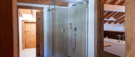A modern shower room with glass doors, a wooden ceiling, and a visible bathtub in the adjacent room.