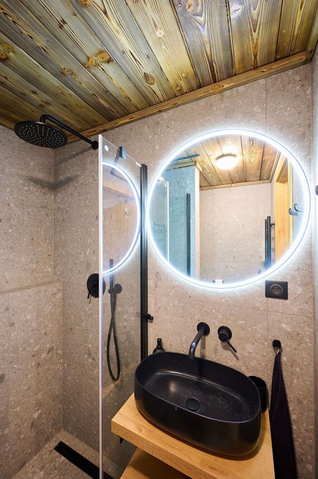 A modern bathroom with a wooden ceiling, a glass-enclosed shower, a round mirror with LED lights, and a black sink.