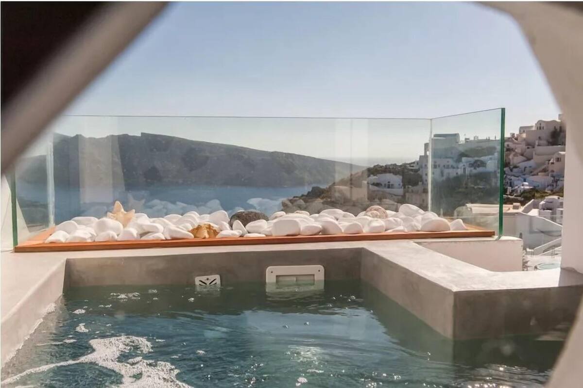 Banyera d'hidromassatge amb pedres blanques i una estrella de mar, envoltada per una barana de vidre, amb vista a una població costanera amb edificis blancs.