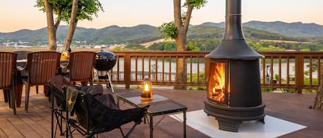 An outdoor deck with a wood-burning stove, wicker chairs, a grill, and a view of a lake and mountains.