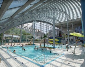 An indoor swimming pool with multiple lanes, a covered area, and a view of buildings outside.