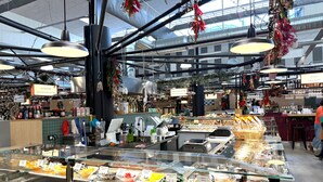 Un mercado con varios puestos de comida, incluyendo una panadería y una charcutería, con decoraciones colgantes y un techo alto.