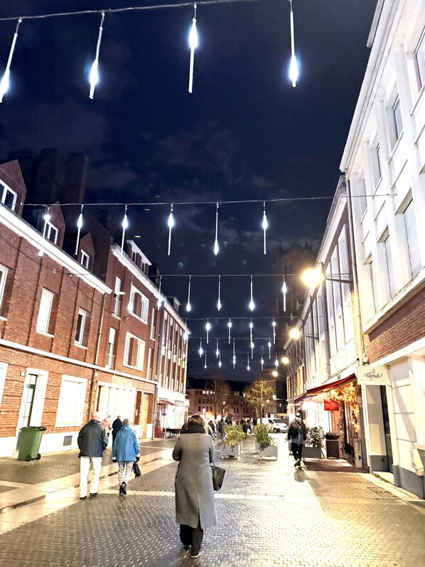 A nighttime street scene with buildings on either side, string lights overhead, and people walking.