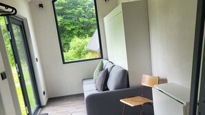 Een modern interieur met een grijze bank, een klein houten tafeltje en een stoel. Een groot raam biedt uitzicht op groen buiten.