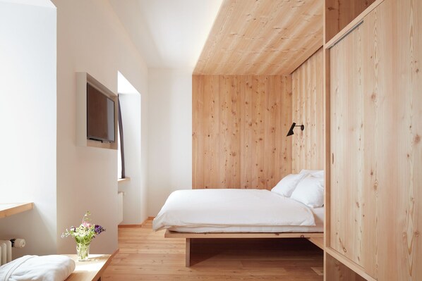 A minimalist bedroom with a wooden bed frame, a bedside table with a vase of flowers, and a wall-mounted TV.