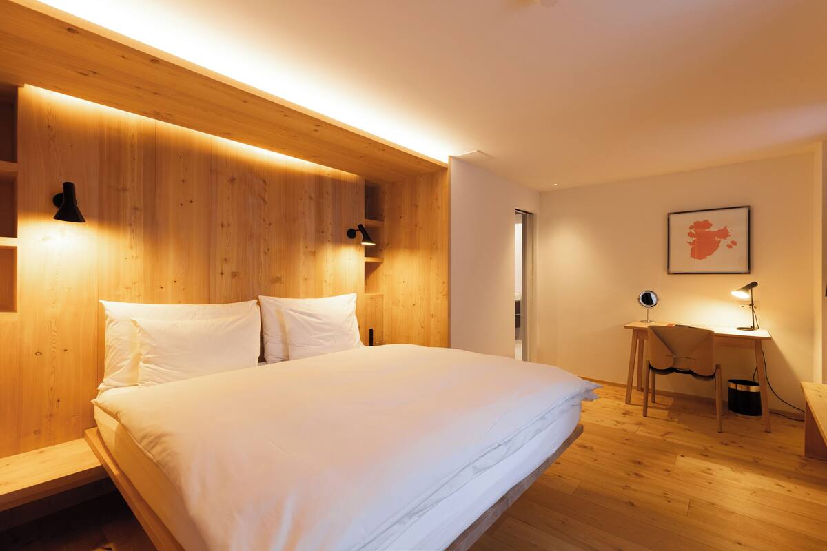 A modern bedroom with a wooden headboard, a bed with white linens, a desk with a chair, and a small trash bin.
