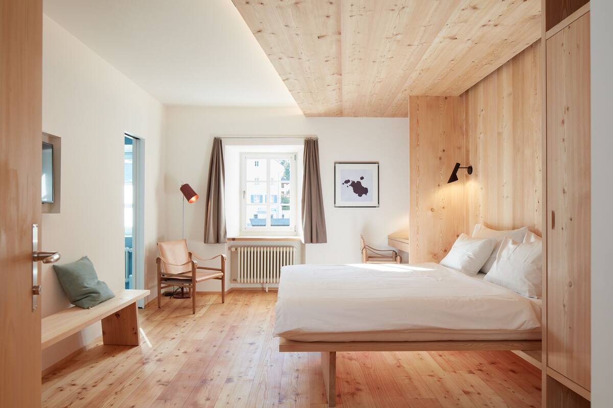 A bedroom with a wooden ceiling, a large bed, a bench, a chair, a window, and a radiator.