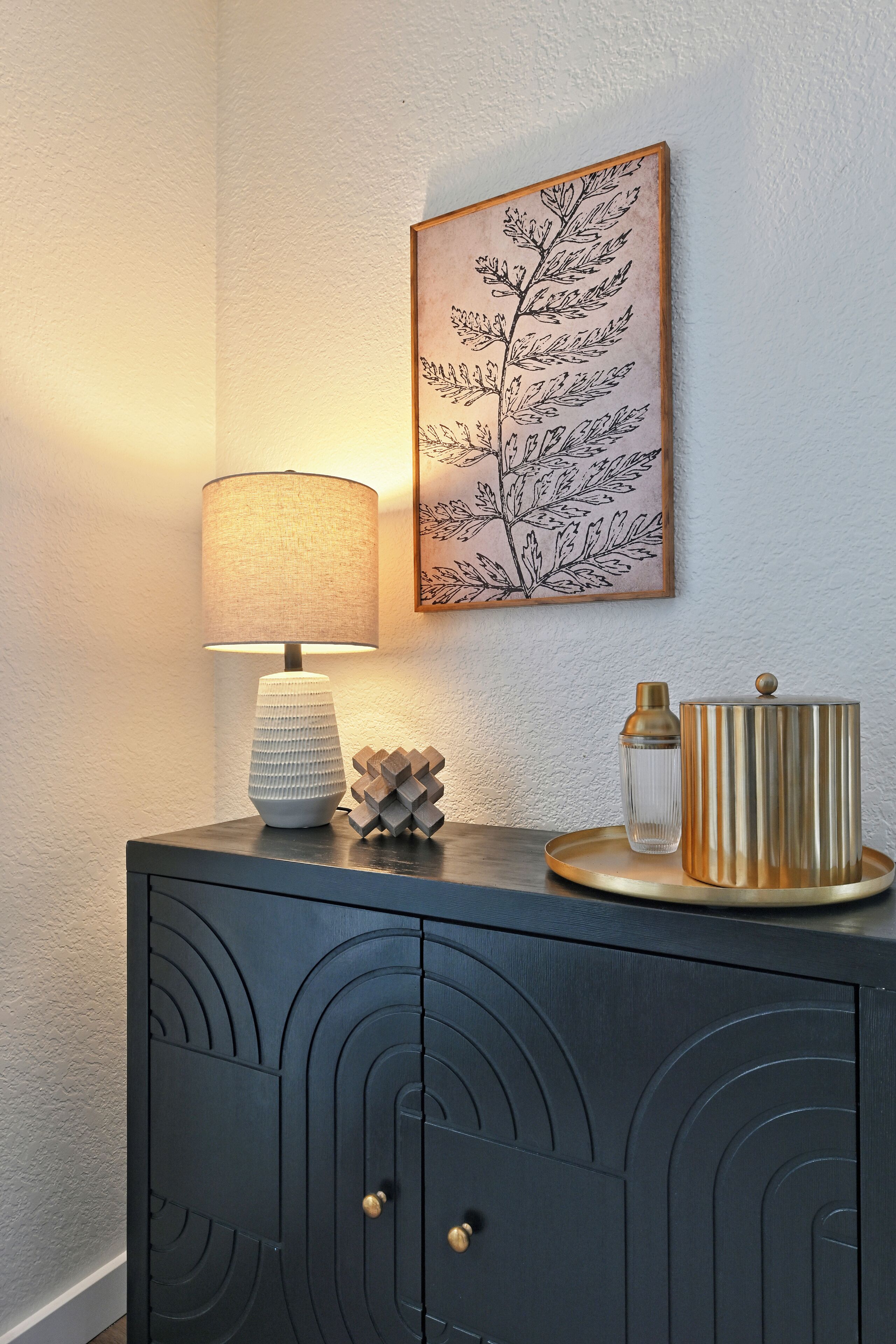 A modern interior with a decorative lamp, a framed botanical print, and a dark cabinet with geometric patterns.