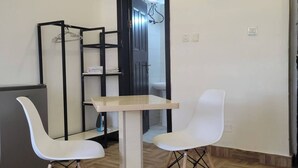 A small, modern room with a wooden herringbone floor, a white table with two white chairs, a black metal shelving unit, and a door leading to a bathroom.