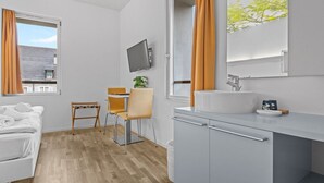 A modern hotel room with a bed, a desk, a chair, a TV, a sink, and a window with curtains.