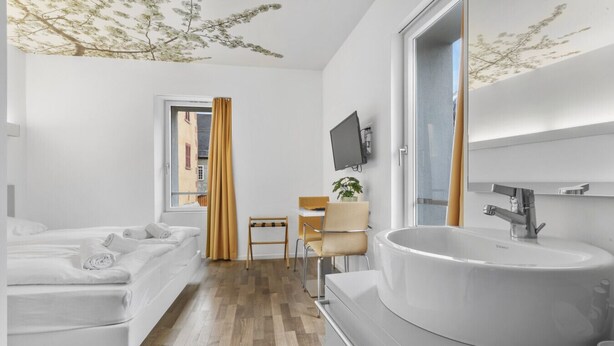A modern hotel room with a large bed, a desk, a chair, a large sink, and a TV mounted on the wall.