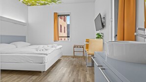 A modern hotel room with a bed, a desk, a chair, a TV, and a bathroom with a sink and mirror.