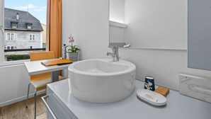 A modern bathroom with a large white sink, a mirror, and a window with a view of buildings. - Cosy single room in the heart of Sierre (Sierre)