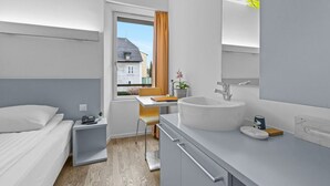 A modern hotel room with a bed, bedside table, desk, chair, and a large bathroom with a sink and mirror. - Cosy single room in the heart of Sierre (Sierre)