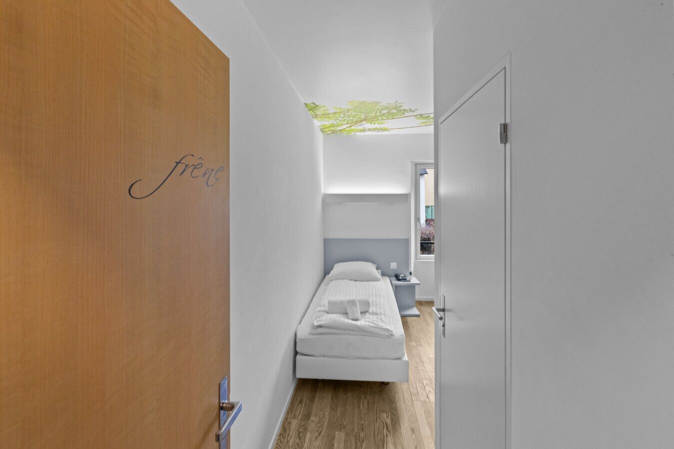 A modern hotel room with a wooden door, a bed with white linens, a nightstand, and a small window.