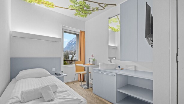 A modern hotel room with a bed, a small desk, a sink, and a mirror. - Cosy single room in the heart of Sierre (Sierre)