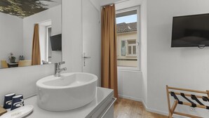 A modern bathroom with a large white sink, a mirror, a wooden rack, and a window with curtains. - Cosy twin room in the heart of Sierre (Sierre)
