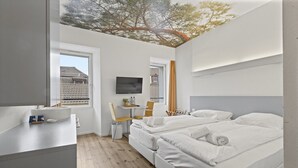 A modern hotel room with two beds, a desk, a TV, and a window with a view of buildings. - Cosy twin room in the heart of Sierre (Sierre)