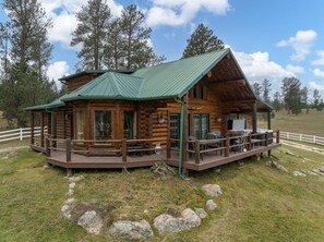 A log cabin with a deck, surrounded by a white fence, set in a grassy area with trees in the background.