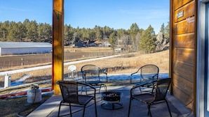 Eine Holzhütte mit Veranda, auf der zwei Stühle und eine Feuerstelle stehen, mit Blick auf eine ländliche Landschaft mit Scheunen und Bäumen.