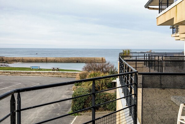 A view of a coastal area with a parking lot, a bench, and a building with a balcony overlooking the sea.