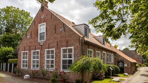 Une maison traditionnelle en briques, dotée d’un toit à pignon abrupt, de nombreuses fenêtres et d’un jardin bien entretenu agrémenté de fleurs et d’arbustes.
