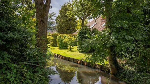 Un jardin paisible, avec un étroit canal d’eau, une végétation luxuriante et une maison en arrière-plan.