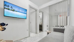 A modern hotel room with a large flat-screen TV displaying a tropical beach scene, a grey sofa, a small round table, and a white-framed sliding door leading to a bathroom. - Premium Jacuzzi Suite at the City Center (Thessaloniki)