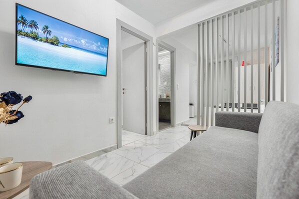 Habitación de hotel moderna con un televisor de pantalla plana grande que muestra una playa tropical, un sofá gris, una mesita redonda pequeña y una puerta corredera con marco blanco que da al baño.