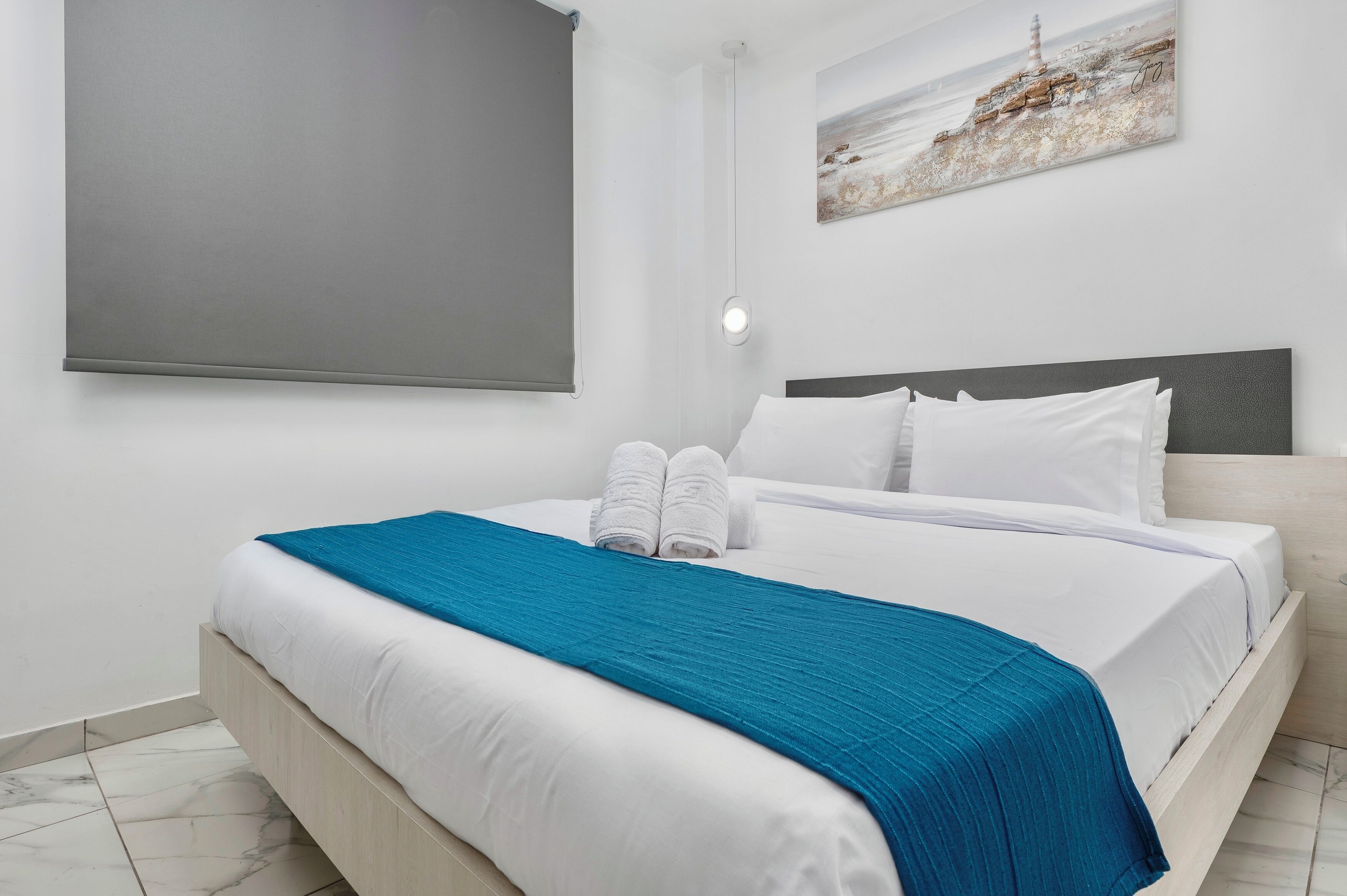 Un lit bien fait, avec du linge de lit blanc agrémenté d’une touche de bleu, une tête de lit et un luminaire fixé au mur.