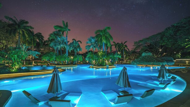 A nighttime pool area with lounge chairs and umbrellas, surrounded by palm trees and illuminated by ambient lighting. - Matapalo 503, beautiful Oceanview Condo with Giant Pool (Tamarindo)