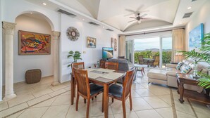 A spacious living area with a dining table, chairs, a sofa, and a TV. There are ceiling fans, a large window with a view, and decorative items on the walls. - Matapalo 503, beautiful Oceanview Condo with Giant Pool (Tamarindo)