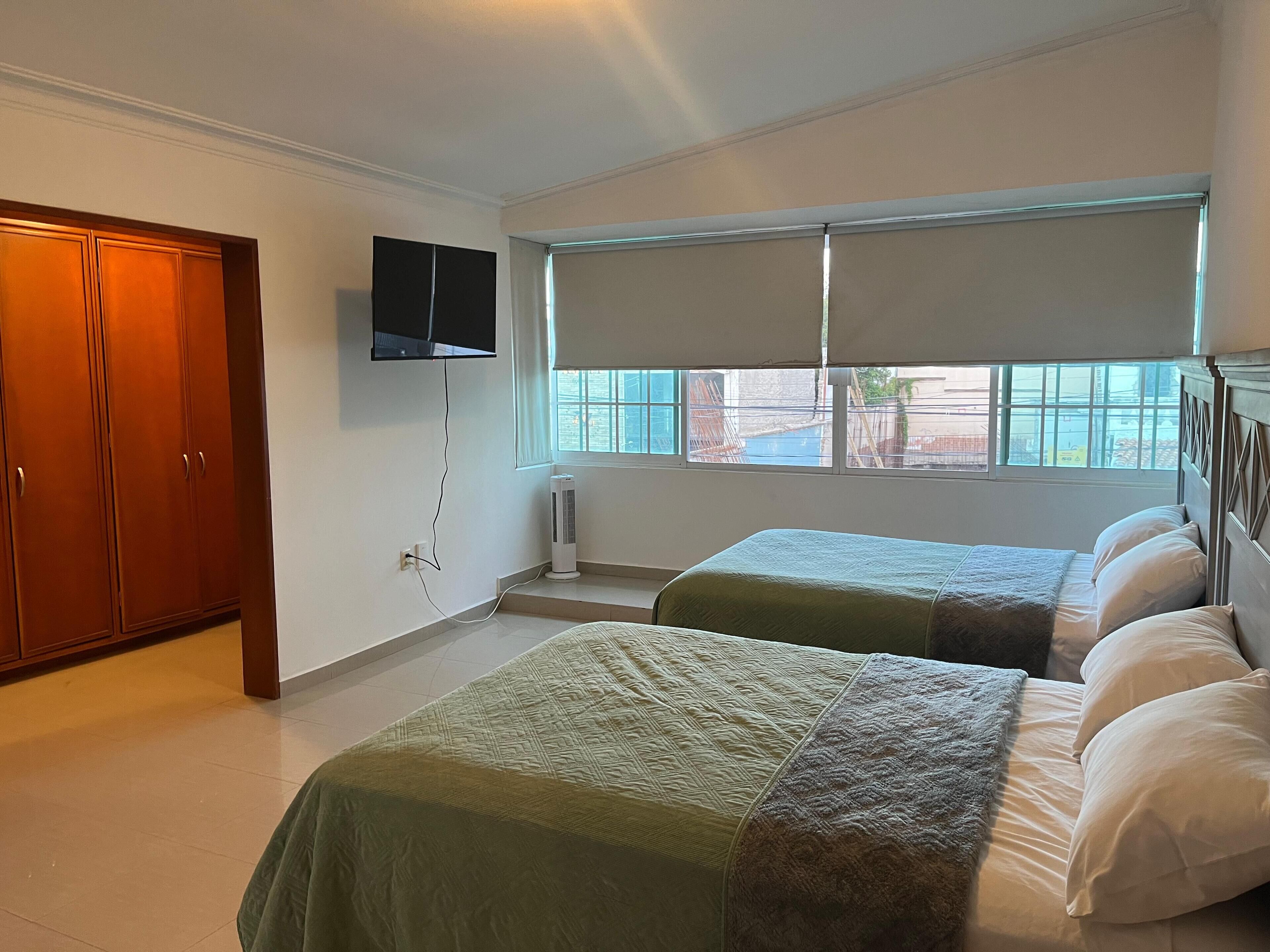 A bedroom with two beds, a TV mounted on the wall, and a window with a view of the outside.