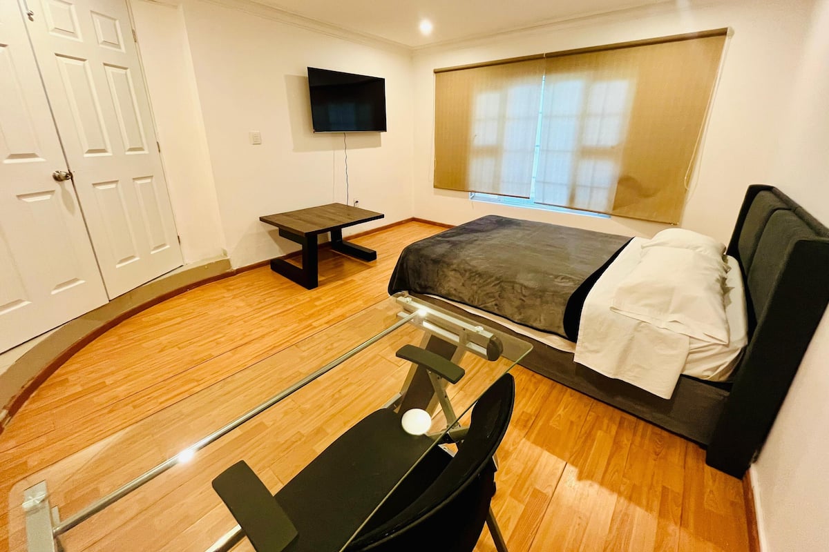 A compact hotel room with a bed, a desk, a chair, a television, and a window with blinds.