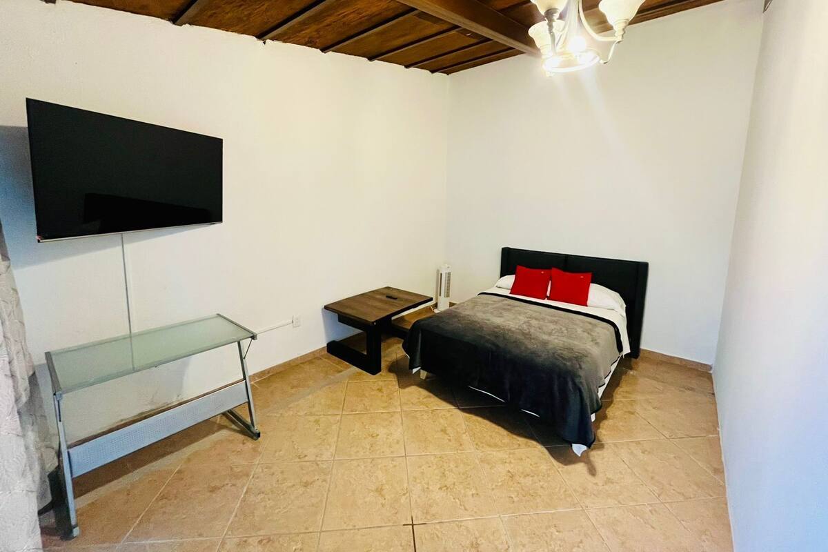 A small, well-lit bedroom with a bed, a TV mounted on the wall, a small table, and a bench.