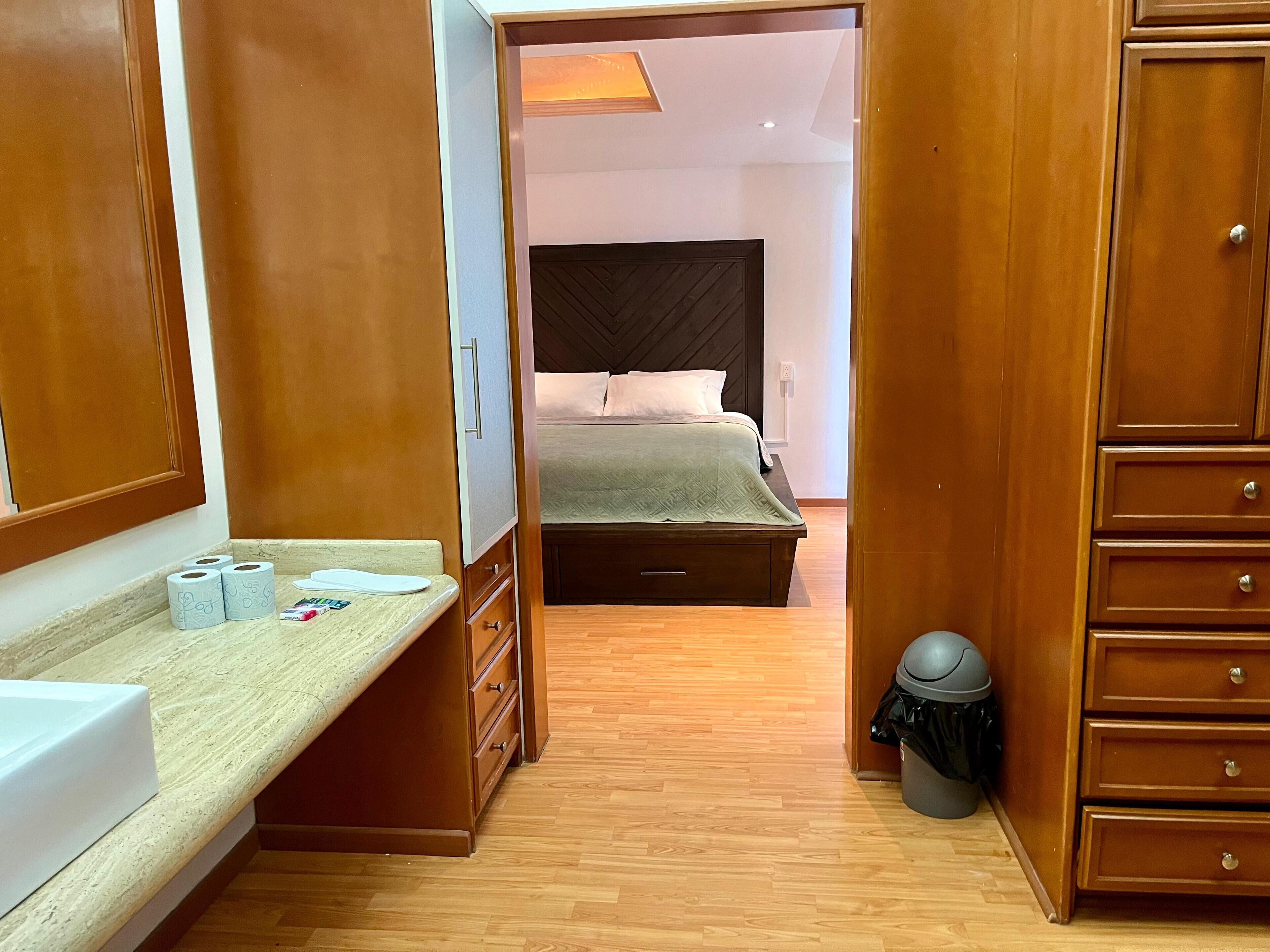 Habitación de hotel con un armario de madera, una cama con cabecera oscura, una mesita de noche con artículos de tocador y un cesto de basura.