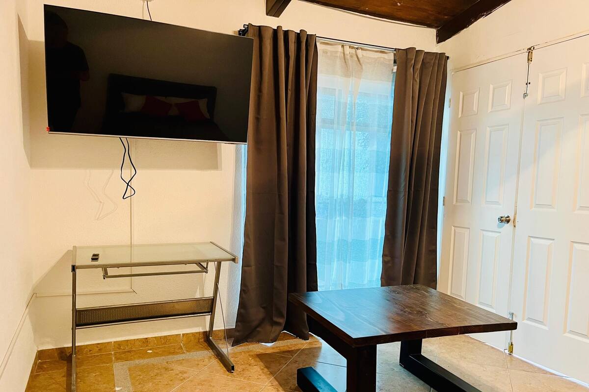 A room with a TV mounted on the wall, a wooden table, a glass shelf unit, and a door with a handle.