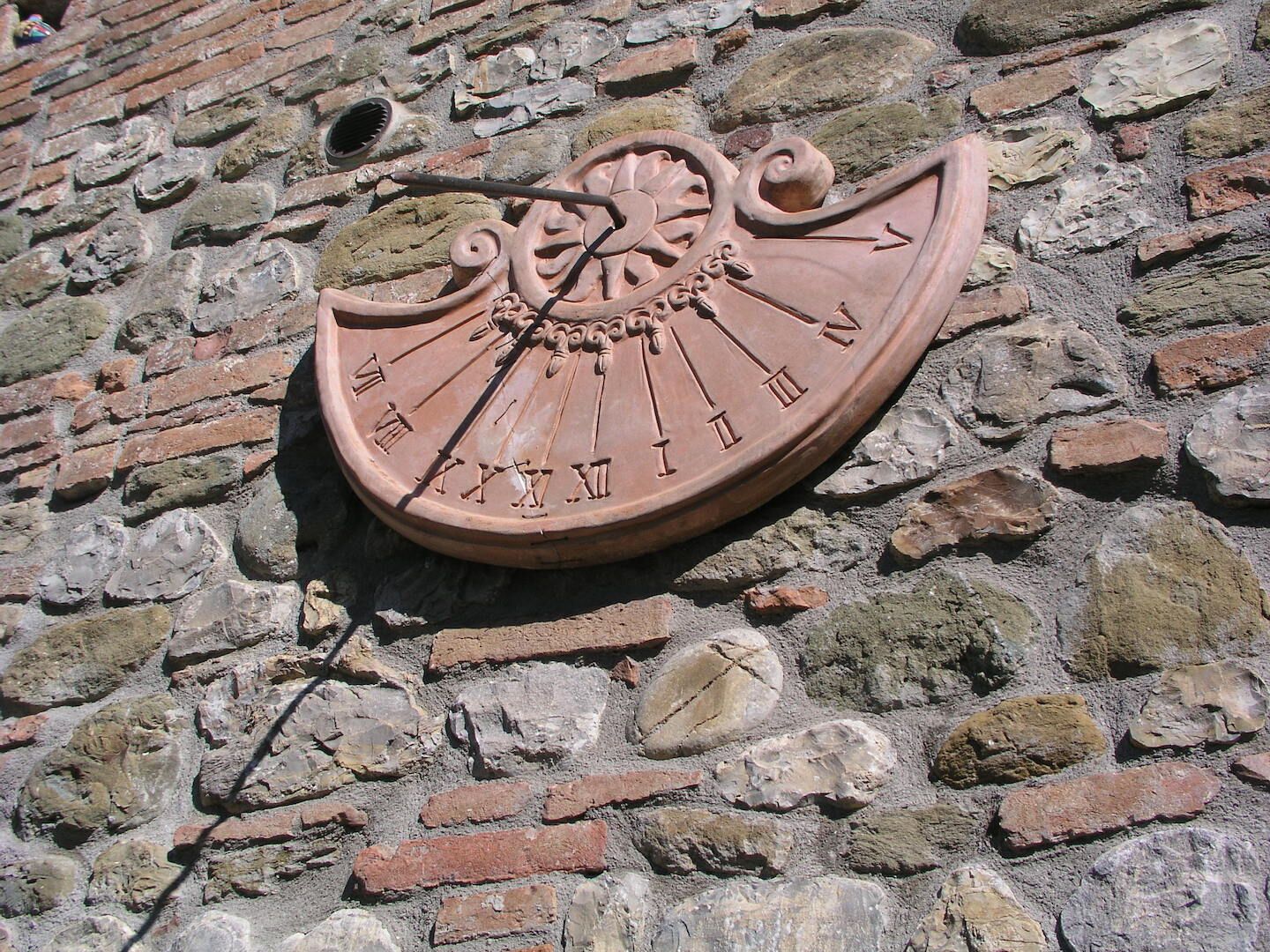 Un cadran solaire orné de chiffres romains et d’éléments décoratifs, fixé sur un mur de pierre.