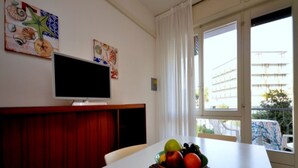 Ein Essbereich mit einem weißen Tisch, einer Obstschale, einem Fernseher an der Wand und einem Fenster mit Vorhängen.