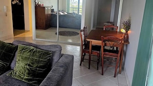 A living room with a grey sofa, a wooden dining table with chairs, and a view through a glass door to a balcony. - Bask in Beach Luxury! (Coolangatta)
