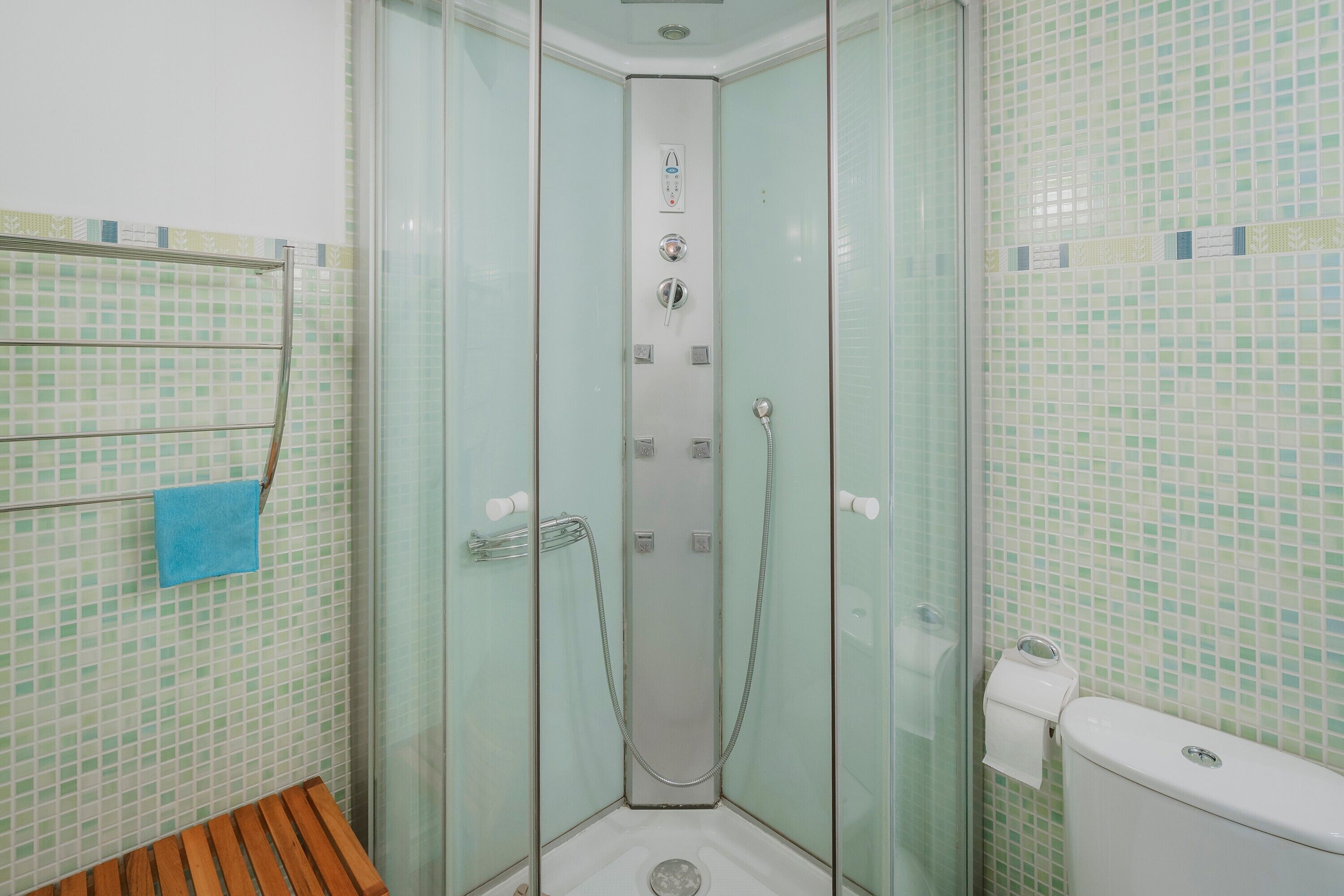 Un box de ducha con paredes de vidrio, un panel de control, un banco de madera y un inodoro con dispensador de toallas de papel.