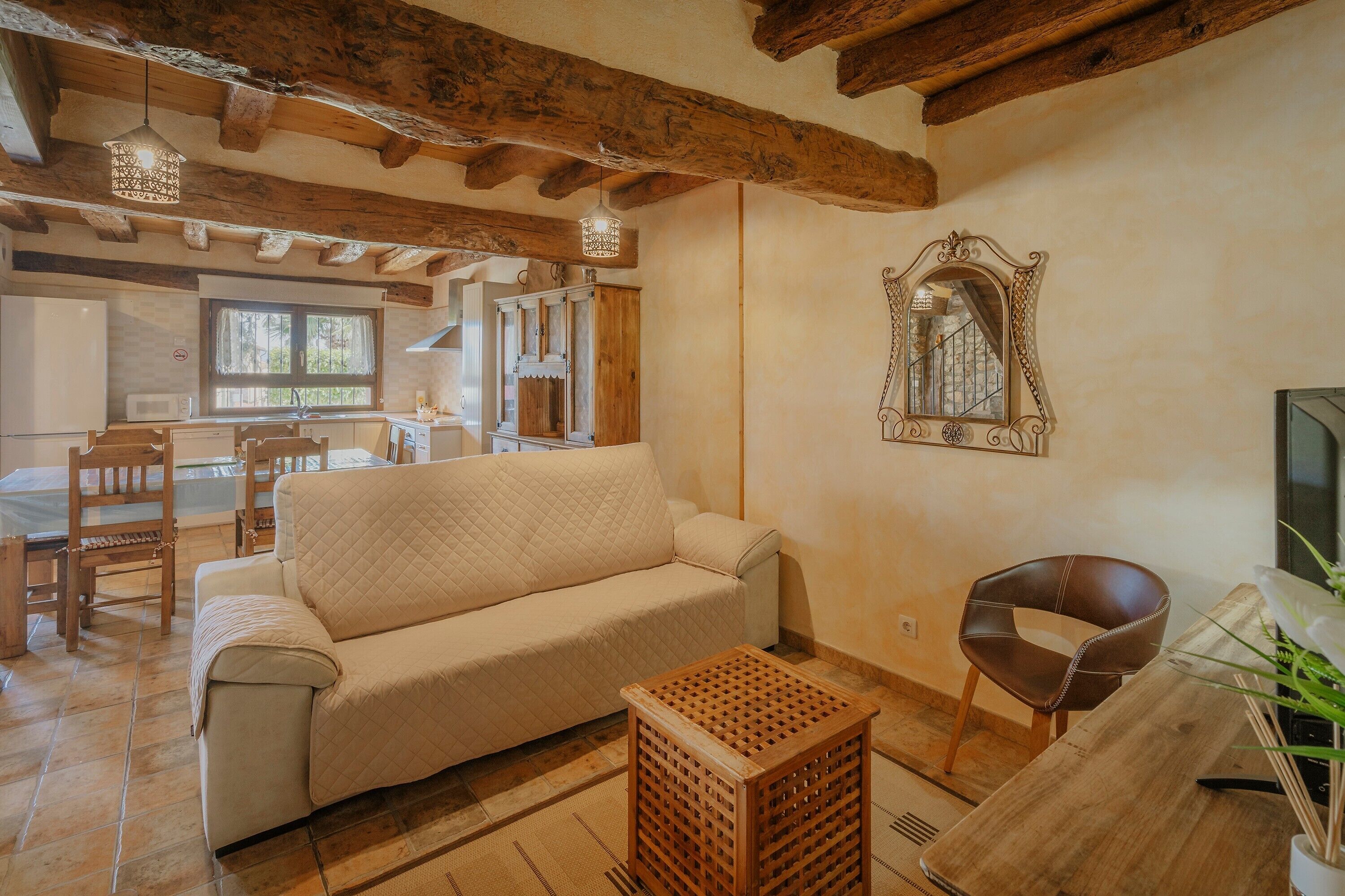 A living room with a sofa, wooden furniture, a mirror, and a kitchen in the background.