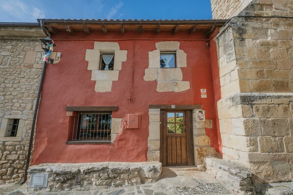 A building with a red facade, stone walls, a wooden door, and a small window with a curtain.