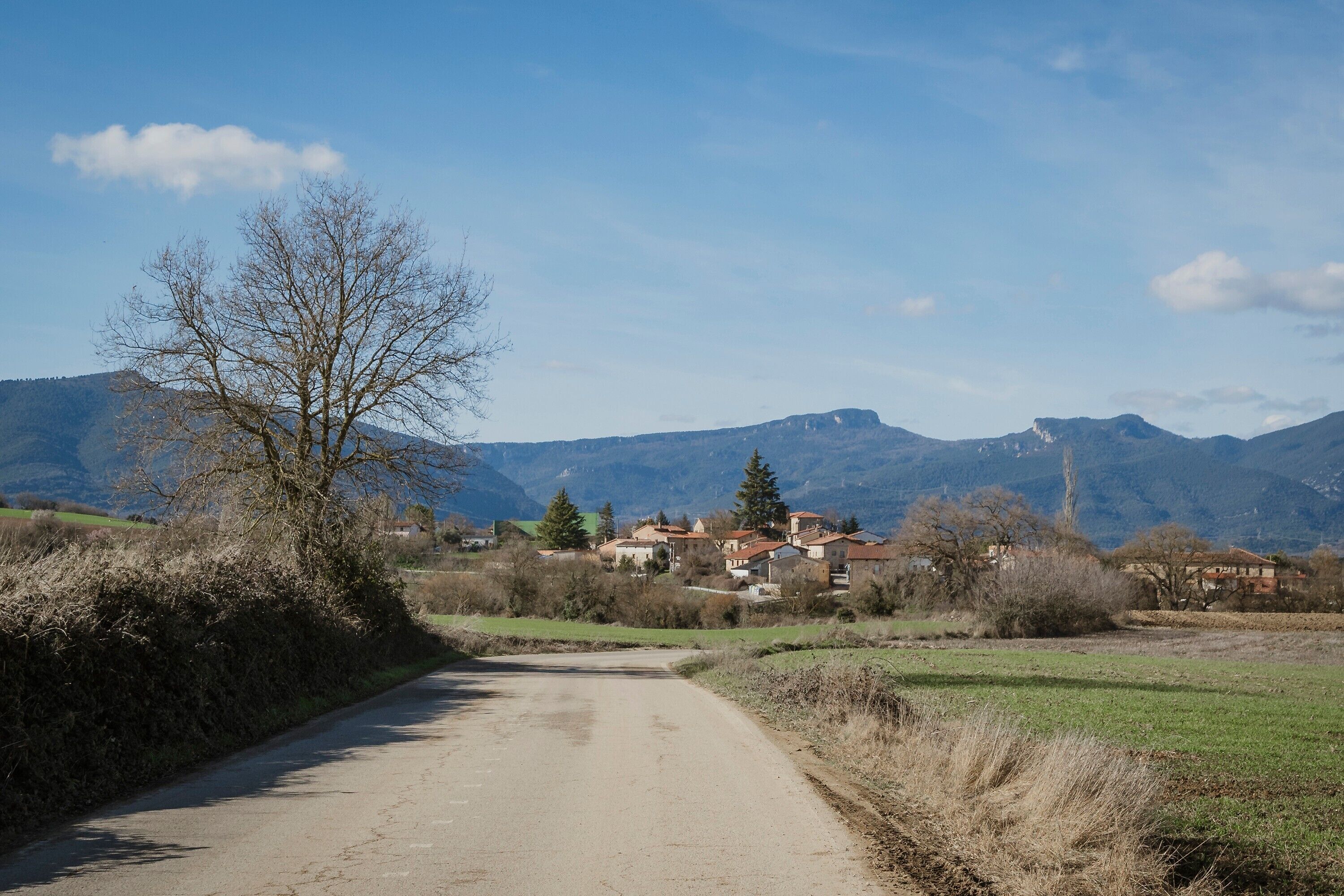 Une route de campagne menant à un village avec des maisons, entouré de montagnes et d’une végétation clairsemée.