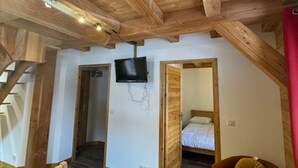 A room with exposed wooden beams, a flat-screen TV mounted on the wall, a bed with a white mattress, and a wooden chair with a cushion.