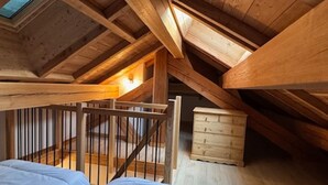 Korkeakattoinen ullakkohuone, jossa on näkyvillä olevat puupalkit, kattovalokuvio ja pieni puinen lipasto.