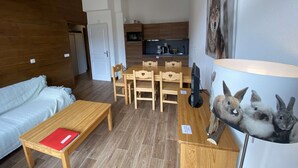 Un salon chaleureux avec une table en bois, un canapé et un coin repas comprenant une table et des chaises en bois. Au fond, on aperçoit une cuisine équipée d’un four à micro-ondes et d’une bouilloire.